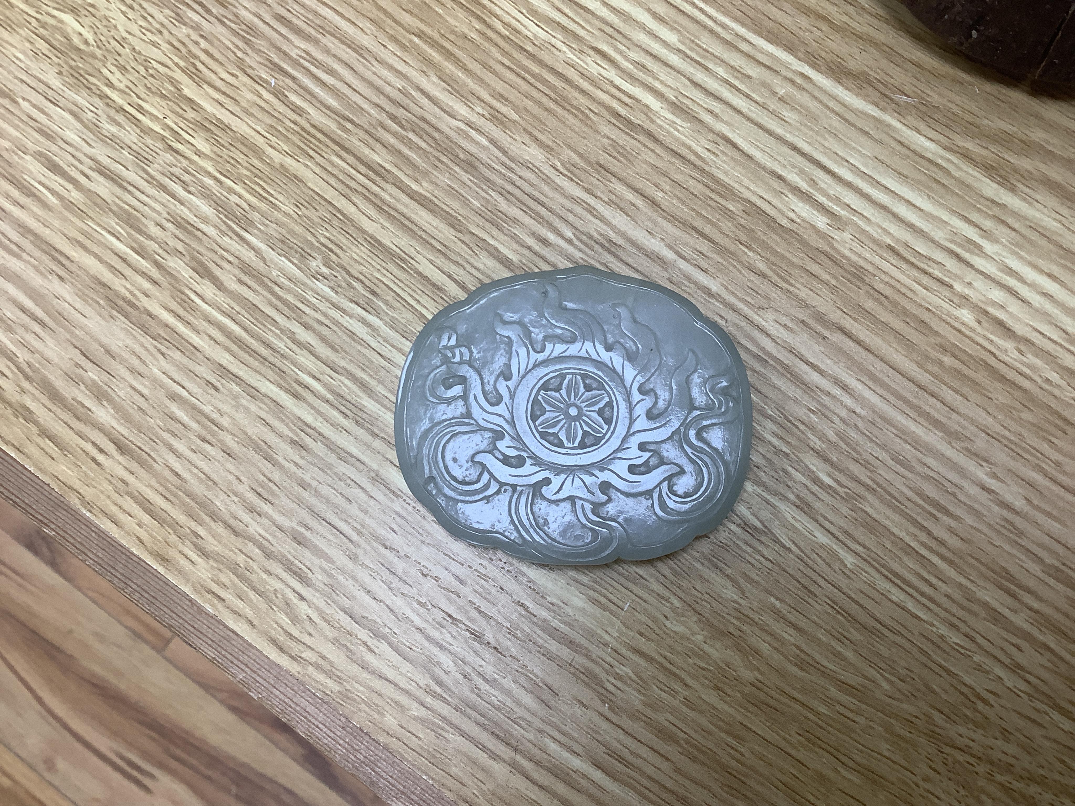 A Chinese pale celadon jade cover for a box, 19th century, carved in relief with a flaming Buddhist wheel emblem, 7.5cm wide. Condition - box base lacking, cover with feint crack